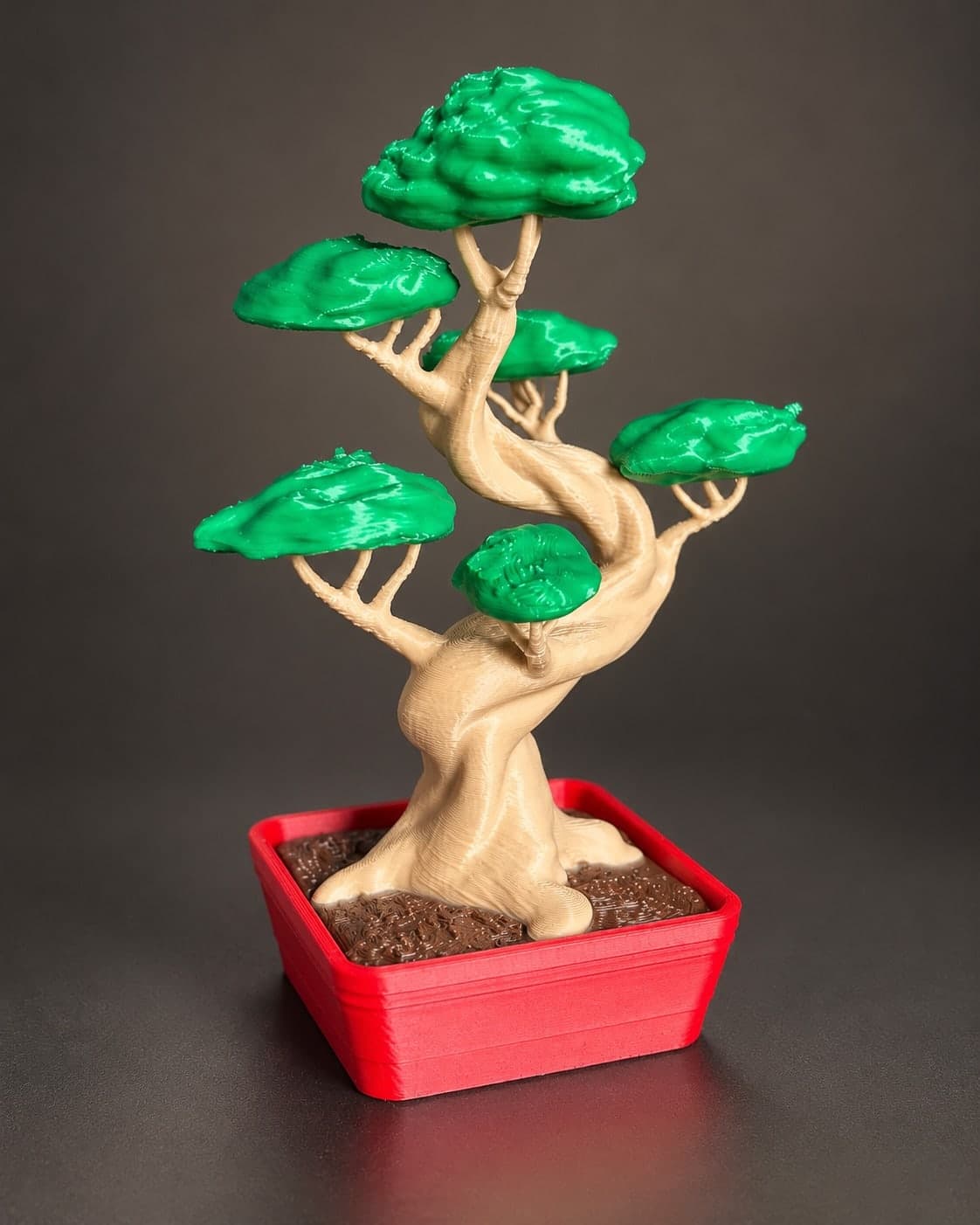 Bonsai impresso em 3D com copa verde, tronco bege retorcido e vaso vermelho, com as impressoras da oficina ao fundo