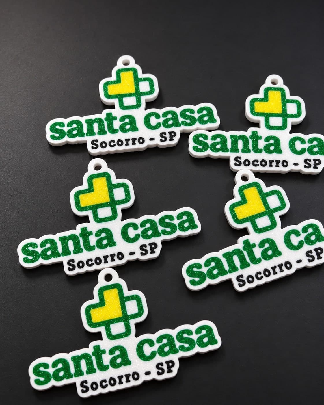 Chaveiros brancos no formato de um logo com cruz verde e amarela e letras verdes, sobre um mousepad