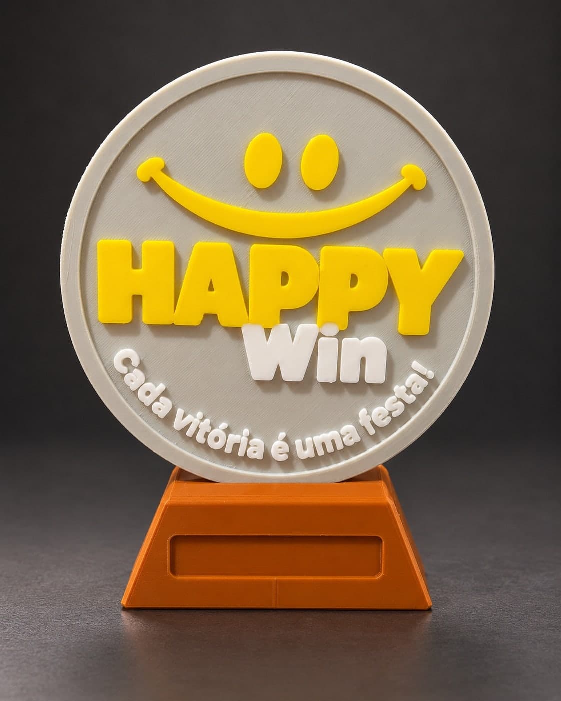 Troféu redondo impresso em 3D com rosto sorridente e letras amarelas e brancas em relevo sobre fundo cinza, em base marrom